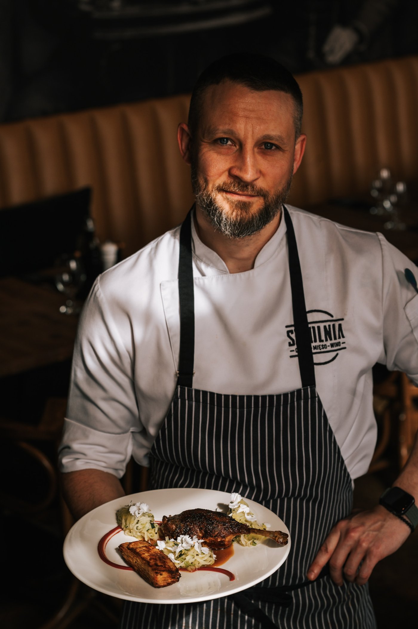 Dawid Golec, head chef of Sztolnia — holding a plate of confit duck inside the restaurant, striped apron over a white chef's jacket with Sztolnia logo